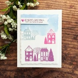 Card with the village houses stamped in soft purple and pink over a pale blue sky panel, deckled edges, sentiment "SEASON'S GREETINGS from our home to yours."