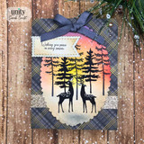 A plaid and navy card with the silhouette buck and doe between tall spruce trees over a sunset blended sky, with a flag reading 'Wishing you peace in every season.'