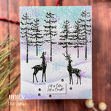 A snowy card with the deer and spruce silhouettes stamped over a lavender and teal winter sky flecked with snow, with a strip reading 'All is Calm, All is Bright.'