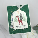 A dark green card with a layered tag of a deer and pine trees stamped in red and green, topped with a gold star and sheer ribbon.