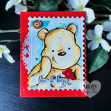 Golden bear holding a red heart on a soft-blue panel inside a bright red scalloped frame, with a script "love & hugs" sentiment.