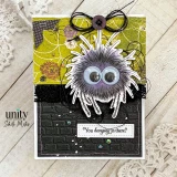 Halloween card with a fuzzy googly-eyed spider hanging over a black brick wall on an olive spiderweb-print background, stamped 'You hanging in there?'