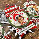 Close up of a Baking Spirits Bright gift tag from Unity Stamp Co: the gingerbread boy in a red Santa hat and gold sunglasses holding a red and white candy cane, on a cream circle inside a green scalloped frame over red and green striped paper and white snowflakes.