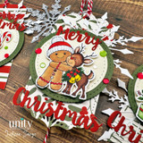 Close up of a Baking Spirits Bright gift tag from Unity Stamp Co: the gingerbread boy in a red and white pom hat hugging a brown reindeer wearing a holly wreath with gold bells, on a cream circle in a green scalloped frame with red Merry Christmas die cuts and white snowflakes.