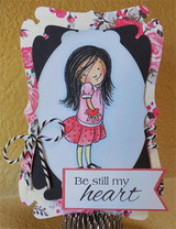 Die cut card with the girl in a pink top and skirt on a pale blue oval framed by floral paper and black trim, tied with black and white twine.