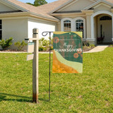 A garden flag featuring a Thanksgiving design with orange pumpkins, displayed on a black metal stand outside a house.