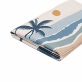 A sandless beach towel featuring blue, tan, and white colours with a palm tree and sun design. It has a logo.