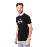 A black short sleeve tee for men featuring a logo on the front. The model is smiling and wearing grey pants.