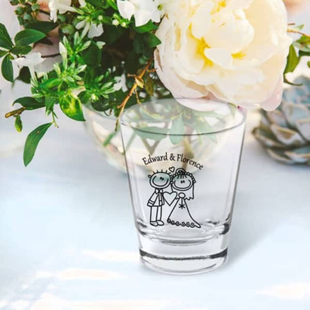 A clear shot glass featuring a couple illustration, surrounded by flowers. The glass has a logo.