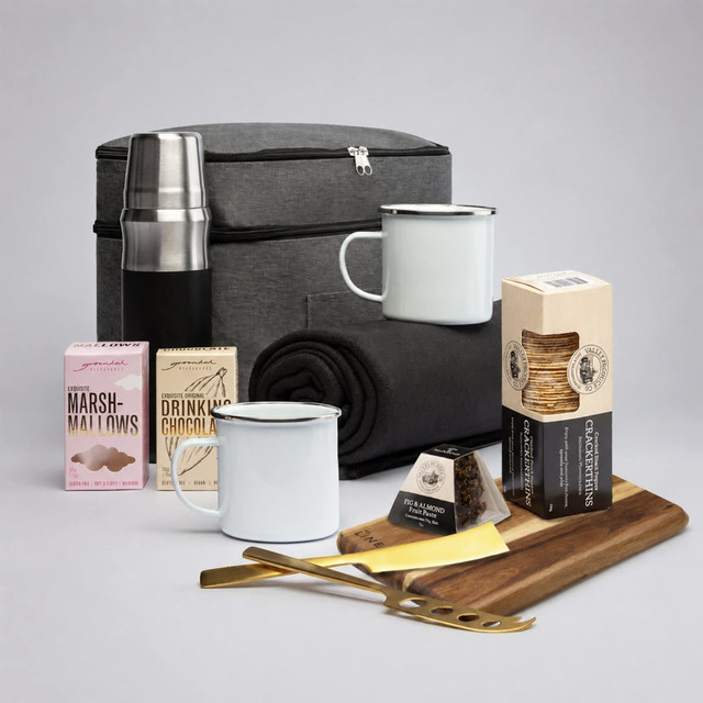 A grey cooler bag with a drink bottle, white mugs, marshmallows, chocolate drink mix, a cheese board, and cheese.