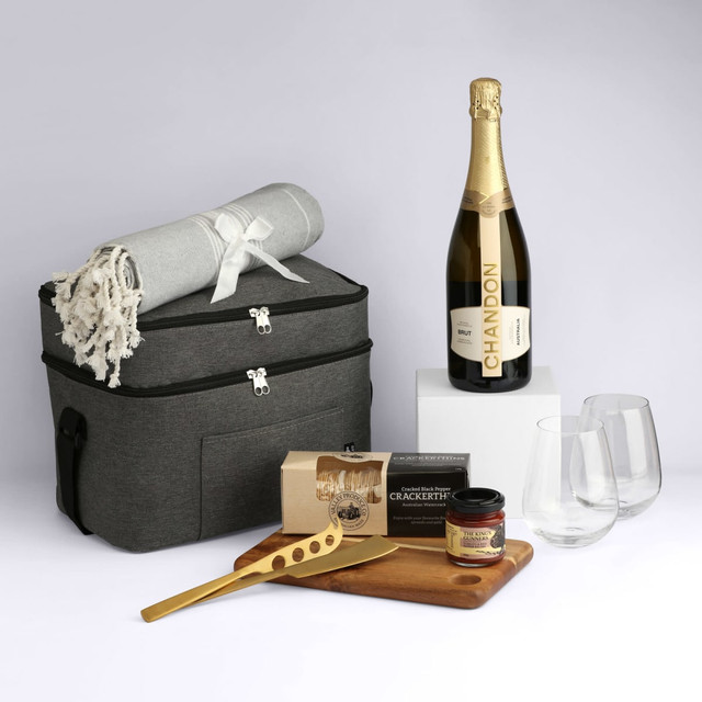 A grey hamper contains a bottle of sparkling wine, two glasses, a cheese board, a blanket, and gourmet snacks.