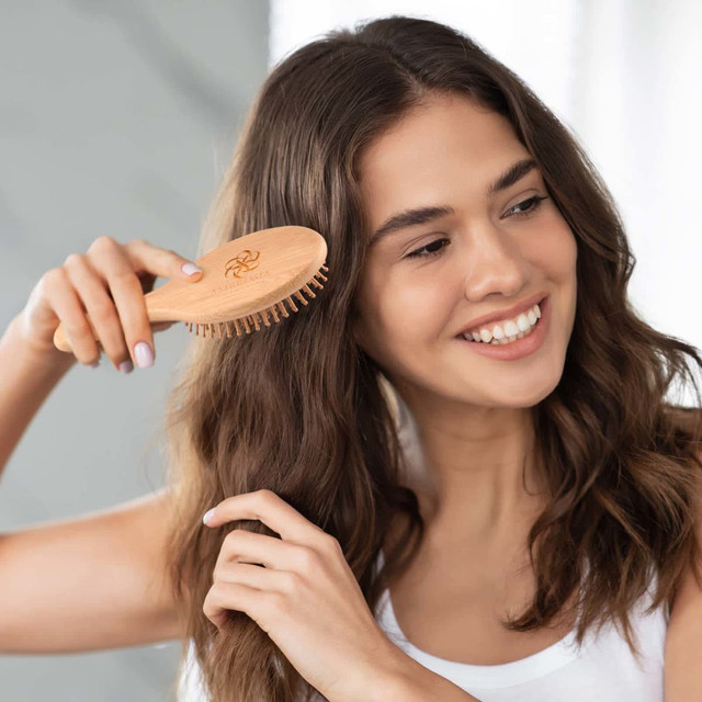 A bamboo hair brush with wooden bristles, held by a woman with wavy, dark hair, smiling in a bright setting.
