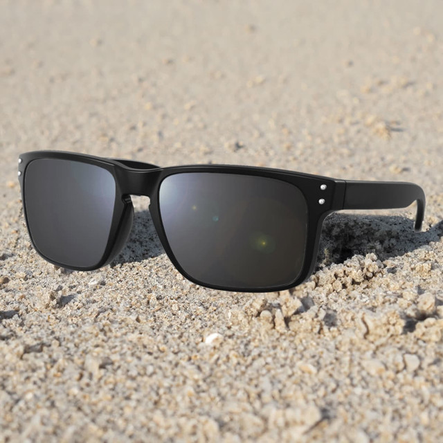 A pair of black sunglasses resting on sandy ground, featuring a simple design and grey lenses.
