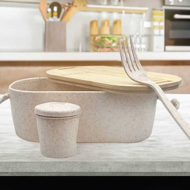 A bamboo lunch box with a light pink hue, accompanied by a small container and a fork, set on a kitchen countertop.