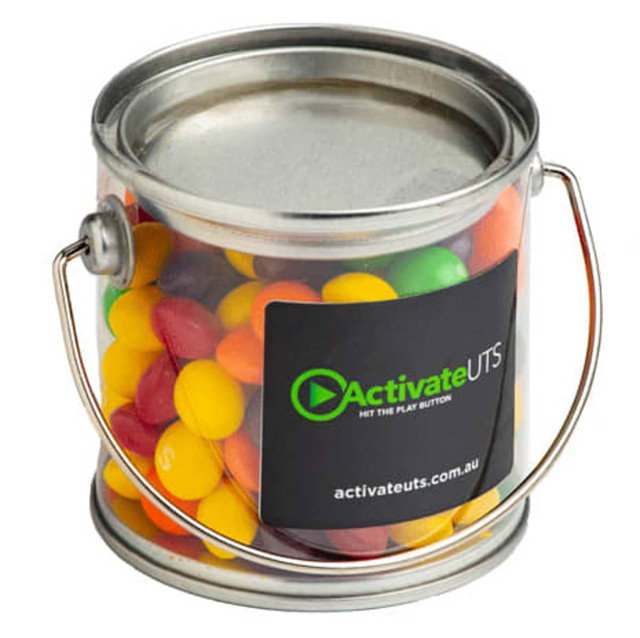 A small clear PVC bucket filled with vibrant Skittles in various colours, featuring a metal handle and a logo.