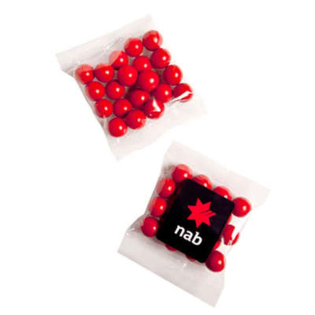 A clear packet containing red chocolate-coated treats and a small black square with a logo.