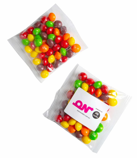 A clear bag containing assorted colourful candy pieces, including red, green, yellow, and orange Skittles. The bag features a logo.