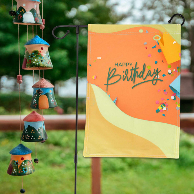 A decorative garden flag featuring a vibrant orange background with colourful confetti and a birthday message.