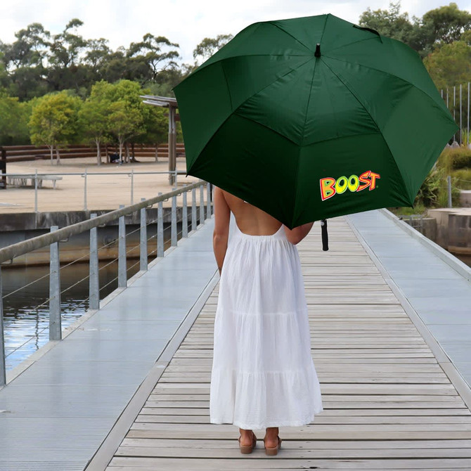 A green umbrella with a logo, held by a person wearing a white dress on a wooden boardwalk near water.