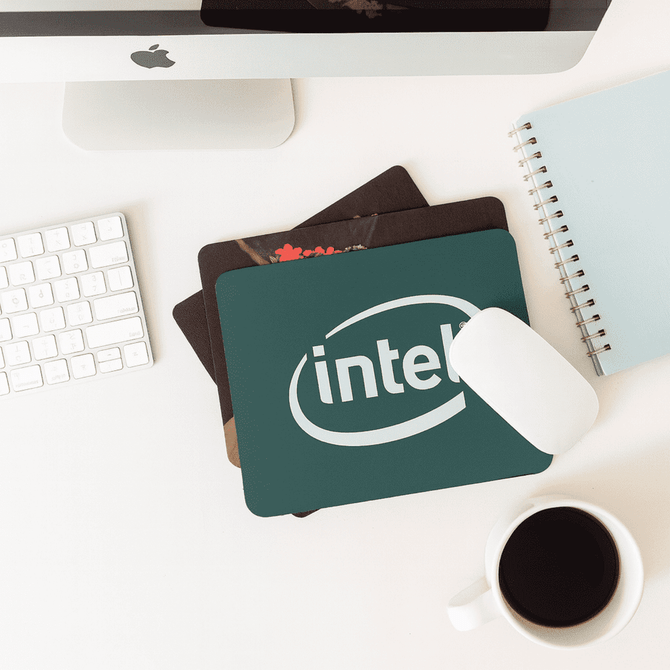 A green fabric mouse mat with a logo, placed on a desk alongside a white mouse, coffee cup, and notebook.