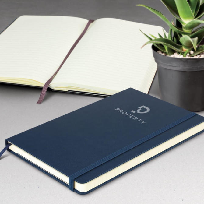 A large, navy blue Moleskine notebook with an elastic closure, featuring lined pages. A potted plant is in the background.