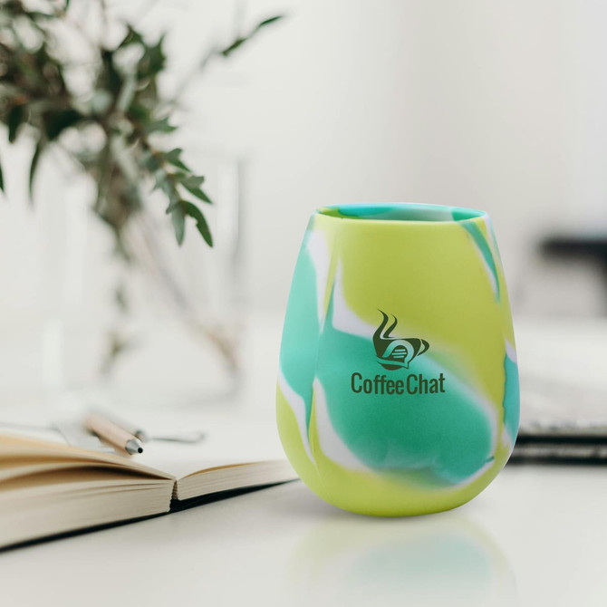 A silicone cup in green and blue marbled design, featuring a logo, on a table with a notebook and a plant.