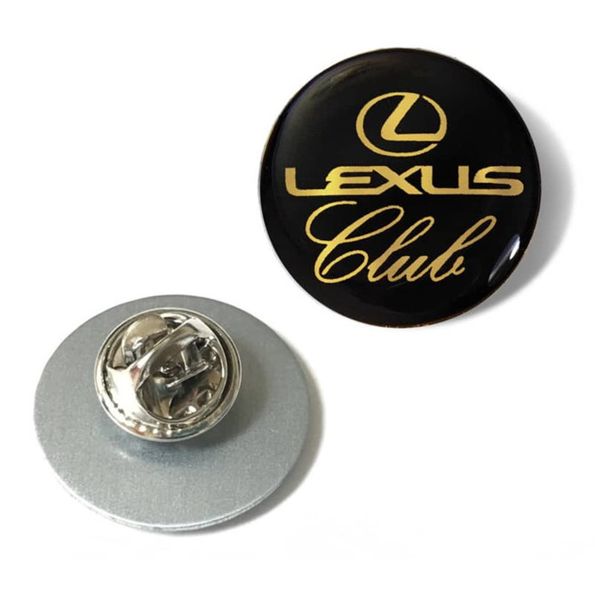 A black lapel pin featuring gold text with a logo and a clasp at the back for attachment.