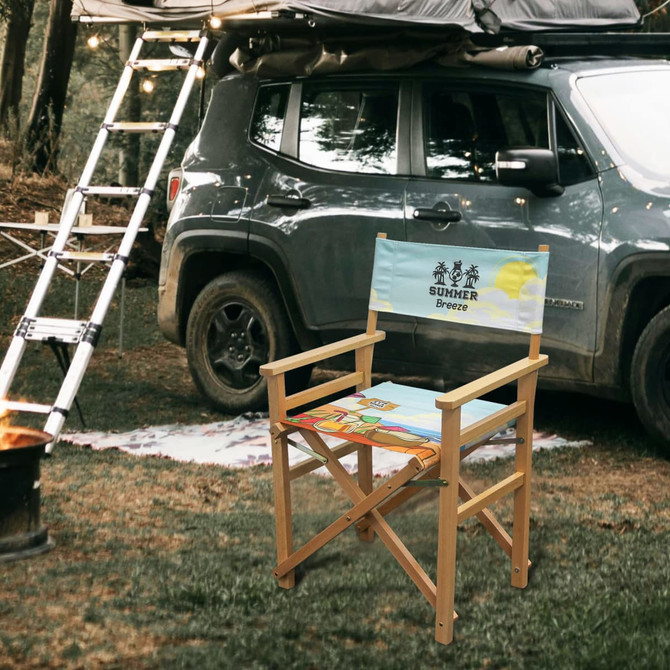A folding director's chair with a light blue seat featuring a beach design, set near a vehicle in a forested area.