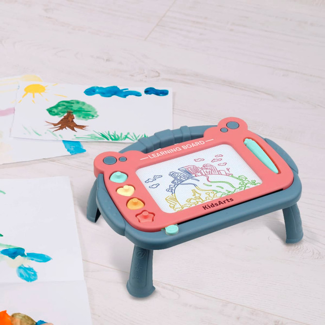 A magnetic drawing board with a stand, featuring grey and pink colours, sits on a light wooden surface beside colourful drawings.