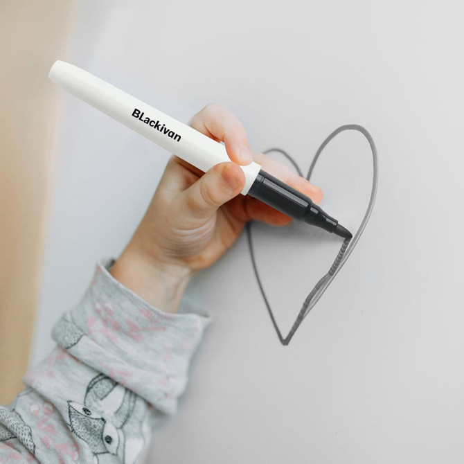 A hand holding a black marker, drawing a heart shape on a light surface. The marker has a white body and a logo.
