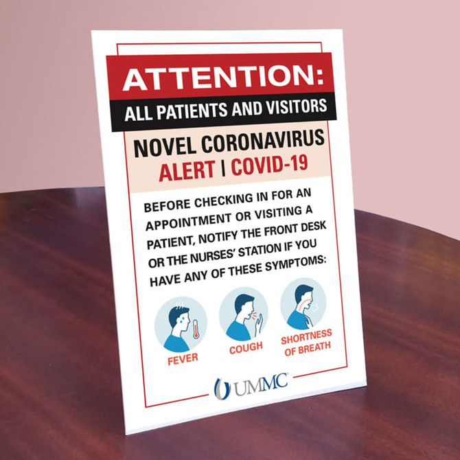 A countertop sign featuring a red and white alert about COVID-19 symptoms with icons for fever, cough, and shortness of breath.