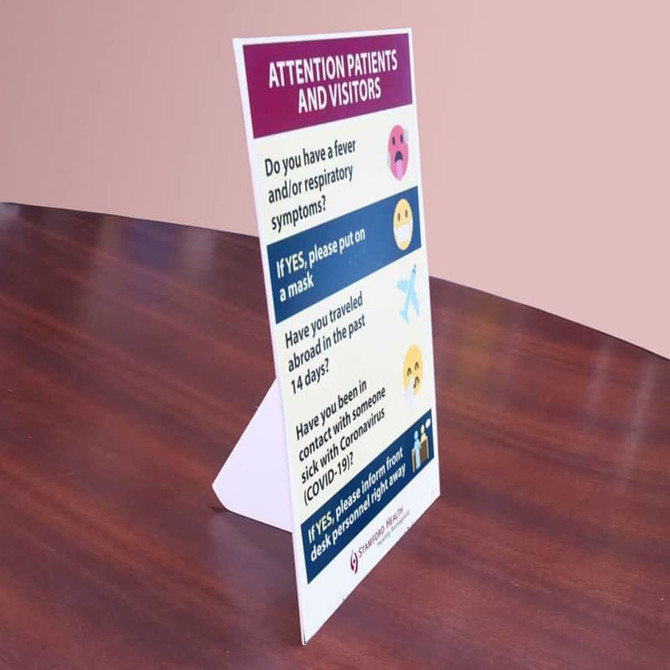 A freestanding sign in white and purple on a wooden table, displaying health check questions for patients and visitors.