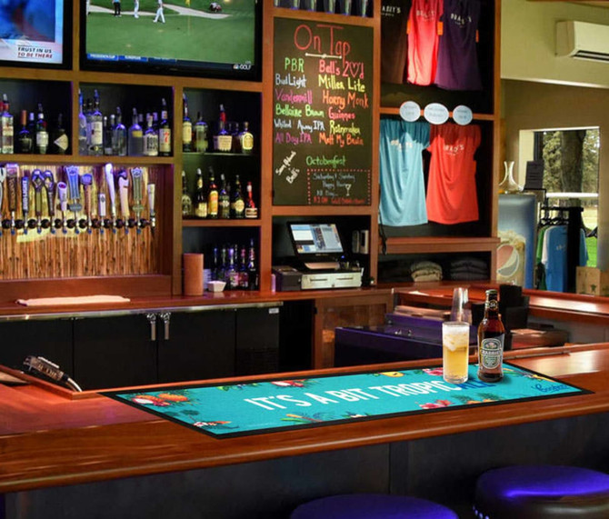 A bar mat featuring a tropical design in vibrant colours, laid out on a bar with drinks nearby.