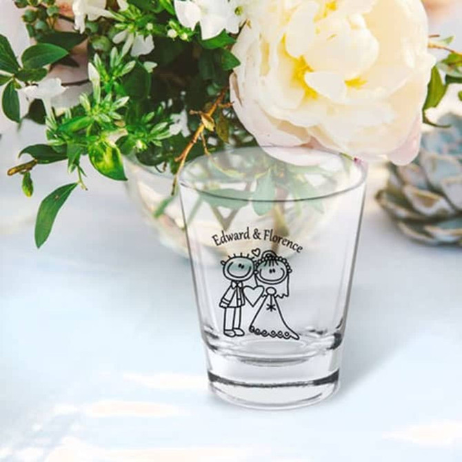 A clear shot glass featuring a couple illustration, surrounded by flowers. The glass has a logo.