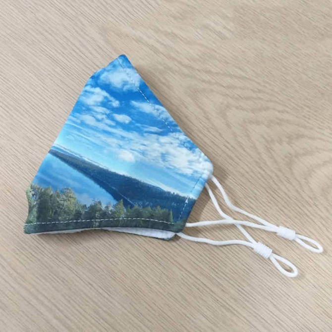 A fabric face mask featuring a scenic blue sky and landscape design with adjustable white ear straps.