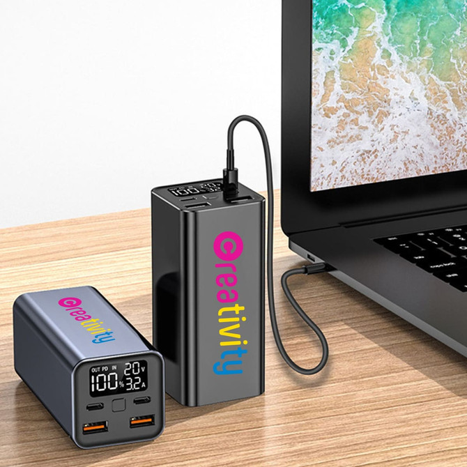 A dark grey power bank with USB ports, connected to a laptop on a wooden surface. The product features a logo.
