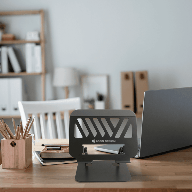 A foldable metal laptop stand in black, displayed on a desk with stationery and a laptop in a modern office setting.