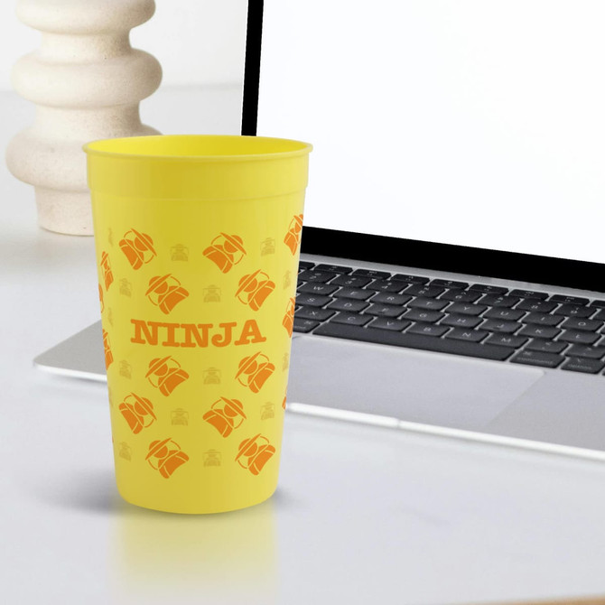 A yellow plastic stadium cup with an orange design and a logo, placed next to a laptop on a desk.