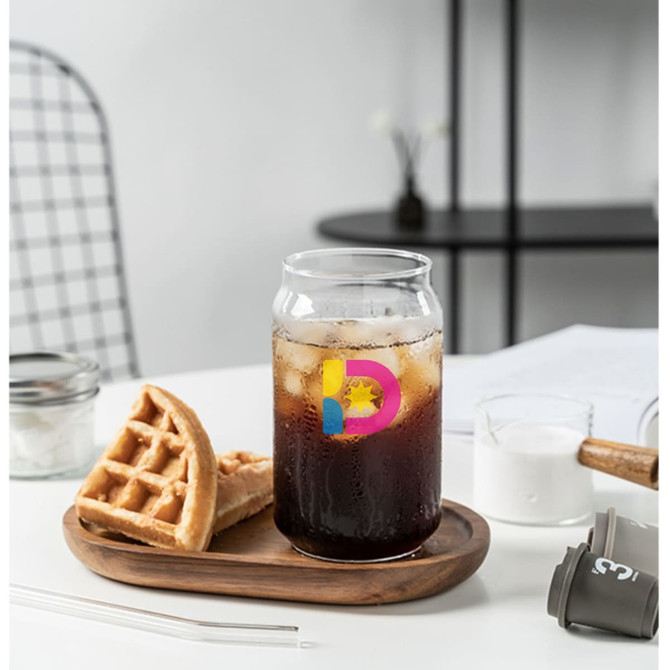A can-shaped glass cup filled with dark fizzy drink sits on a wooden tray beside a waffle. The cup features a logo.