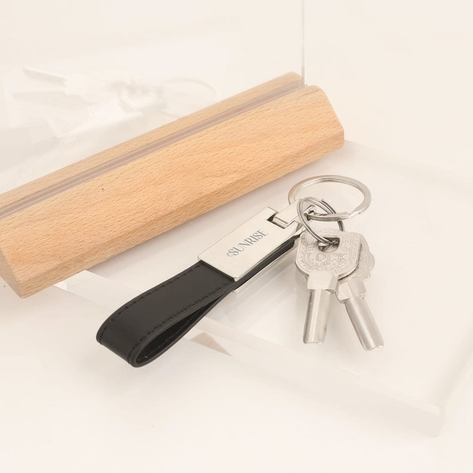 A PU leather keyring in black with a metal tag, holding two silver keys and displayed with a wooden block.