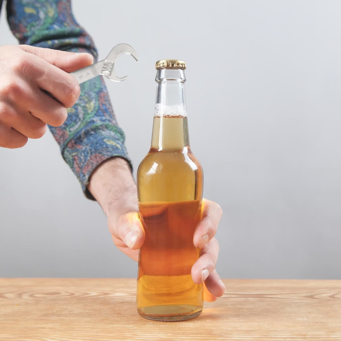 A drink bottle opener shaped like a wrench in silver, held by a hand over a beer bottle on a wooden surface.