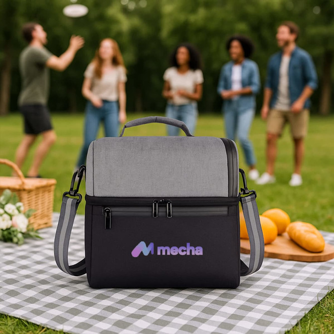 A two-compartment lunch bag in grey and black with a logo, resting on a picnic blanket. People are enjoying a picnic in the background.