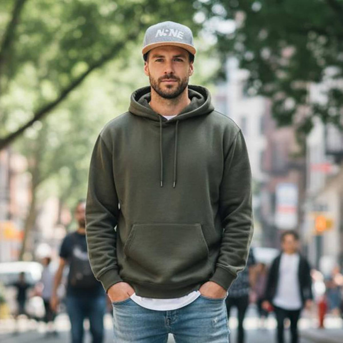 A man in an olive green hoodie and a grey snapback cap with a logo, standing on a busy street surrounded by people.