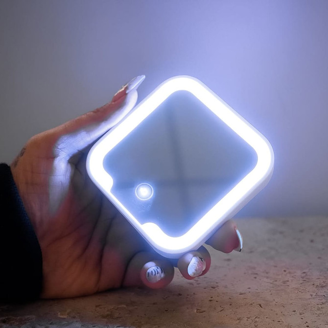 A compact mirror with an LED light, featuring a square shape and held in a hand. The frame glows softly in white.