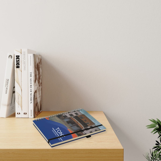 A5 wiro side bound notebook in blue, placed beside stacked books on a wooden surface with a plant in the background.