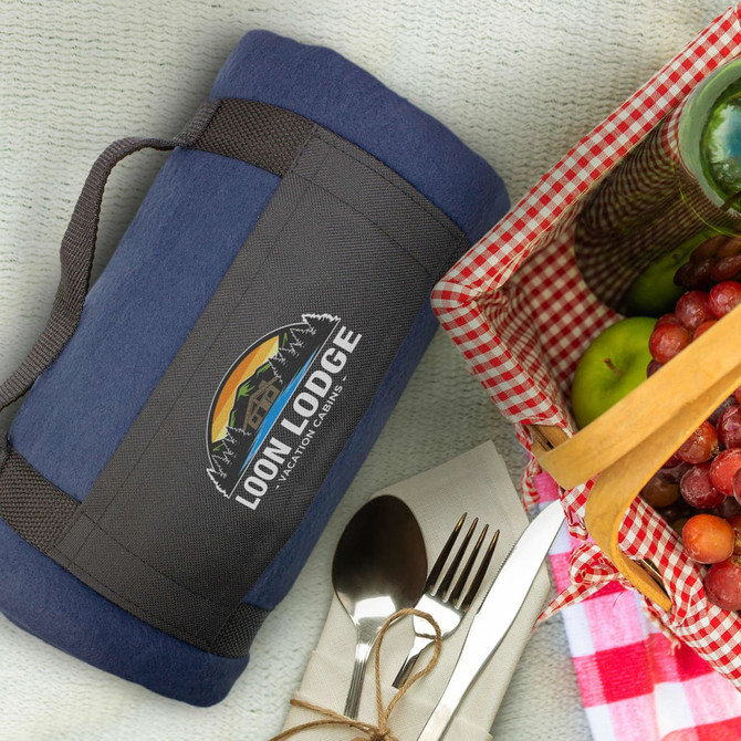 A navy fleece blanket with a strap, featuring a logo, next to a picnic basket and cutlery.