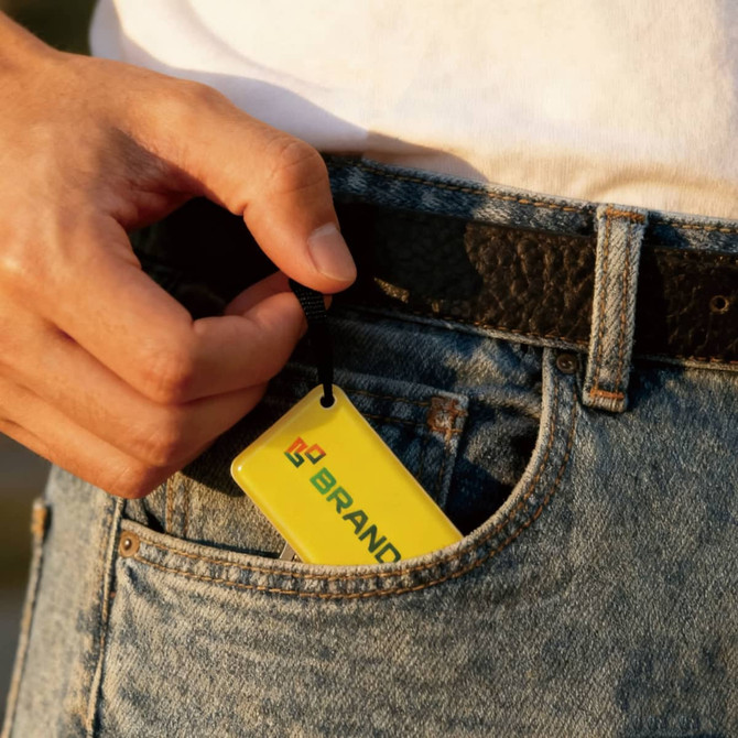 A hand holding a yellow NFC epoxy square tag, with a logo, tucked into the pocket of denim jeans.