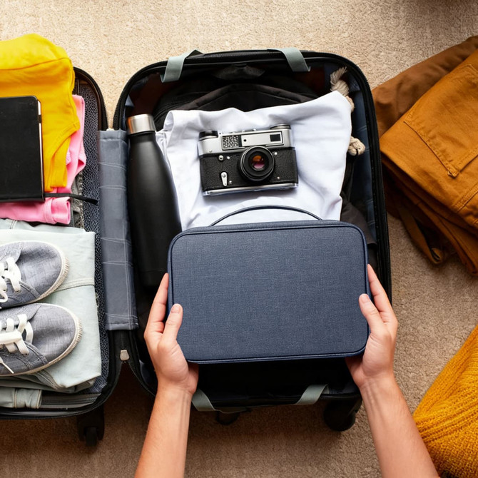 A navy portable hanging travel toiletry bag being held above a suitcase filled with clothing, a drink bottle, and a camera.
