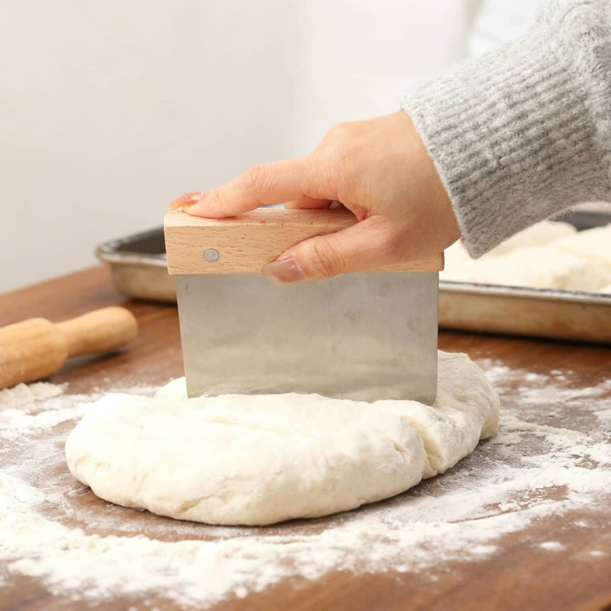 A beechwood handle flour cutter with a metal blade is shown cutting dough on a wooden surface, surrounded by flour.