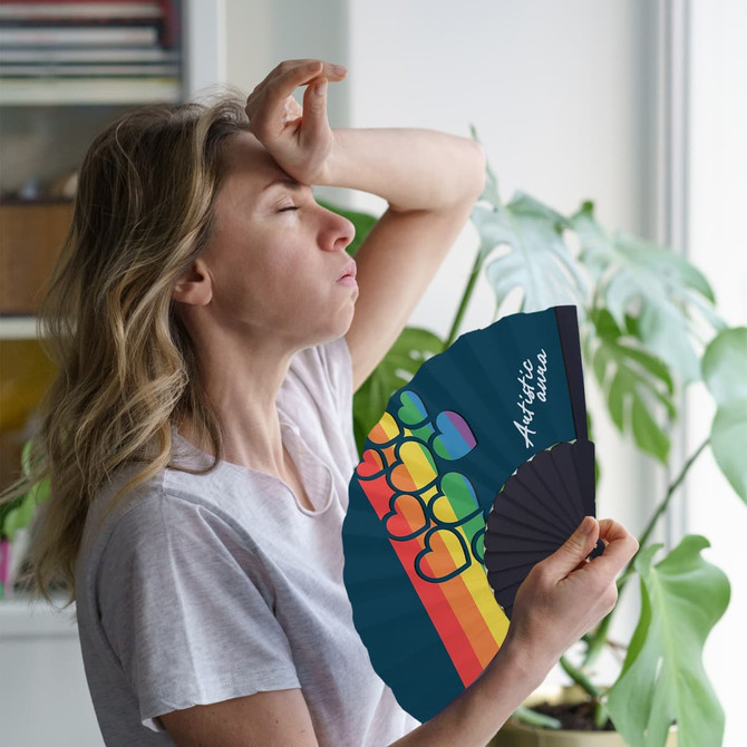 A paper hand-held fan with a black interior and a colourful colourful heart design. A woman uses it while looking warm.